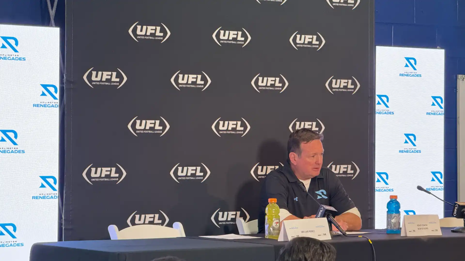 Coach Bob Stoops giving pressor after Renegades game. Behind him is a black step and repeat pattern.