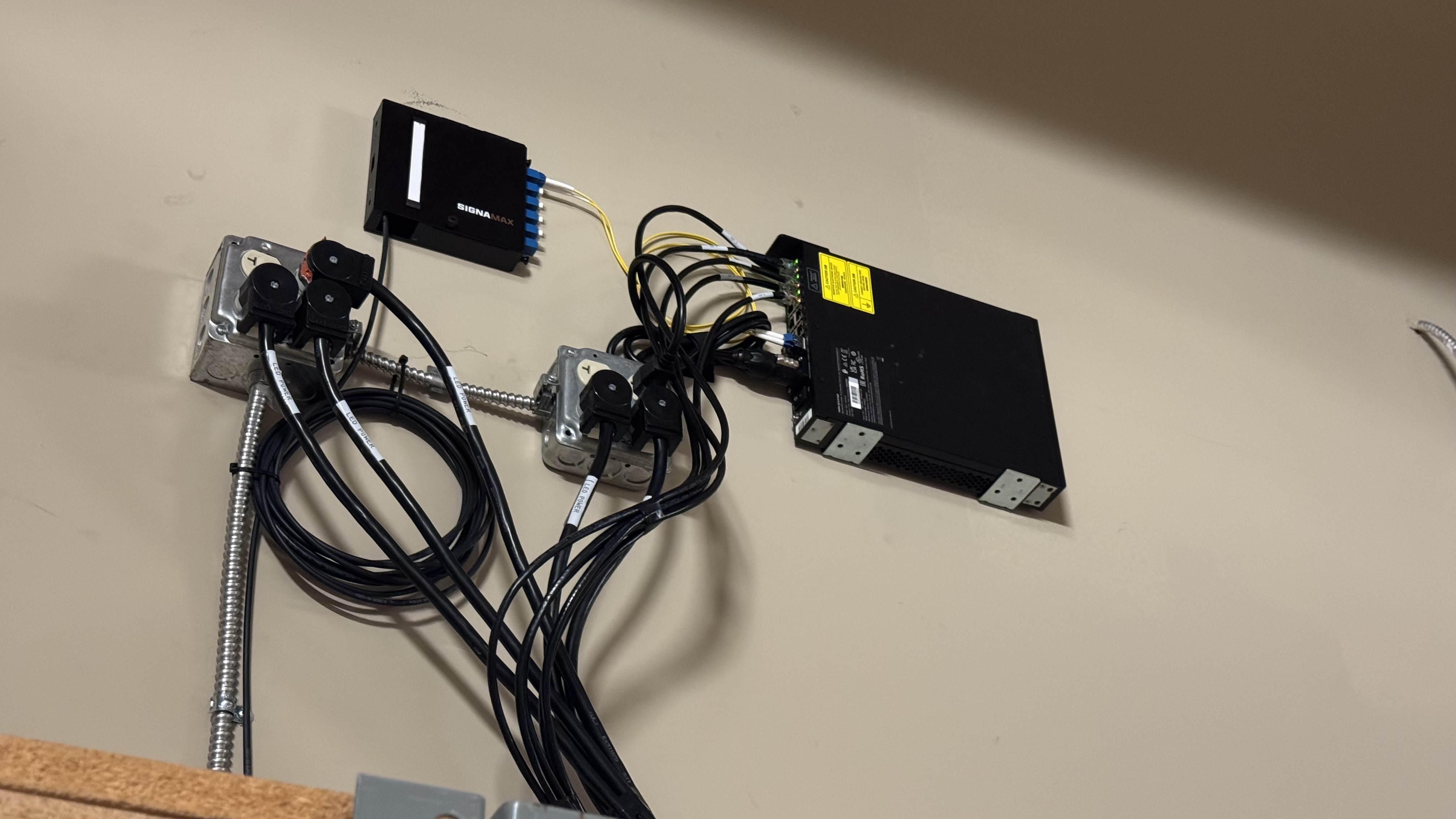 A close-up of a fiber optic cable conversion setup on a wall, featuring a SIGNA MAX switch, power outlets, and neatly arranged wiring.