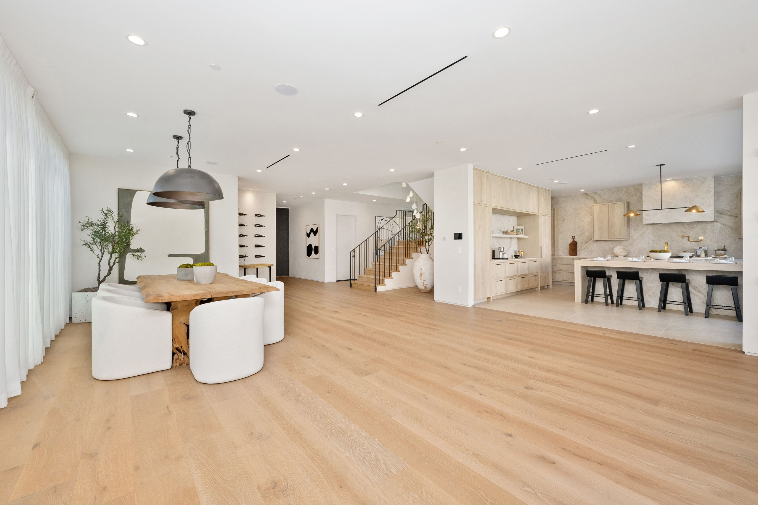 Open-concept modern living space featuring a light wood dining table with white upholstered chairs, minimalist kitchen with bar stools, and light wood flooring throughout.