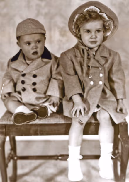 Black and white photo of two young children sitting on a bench, dressed in vintage coats and hats.