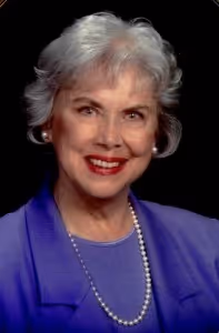 Smiling older woman with short gray hair wearing a purple blazer, pearl necklace, and earrings against a black background.