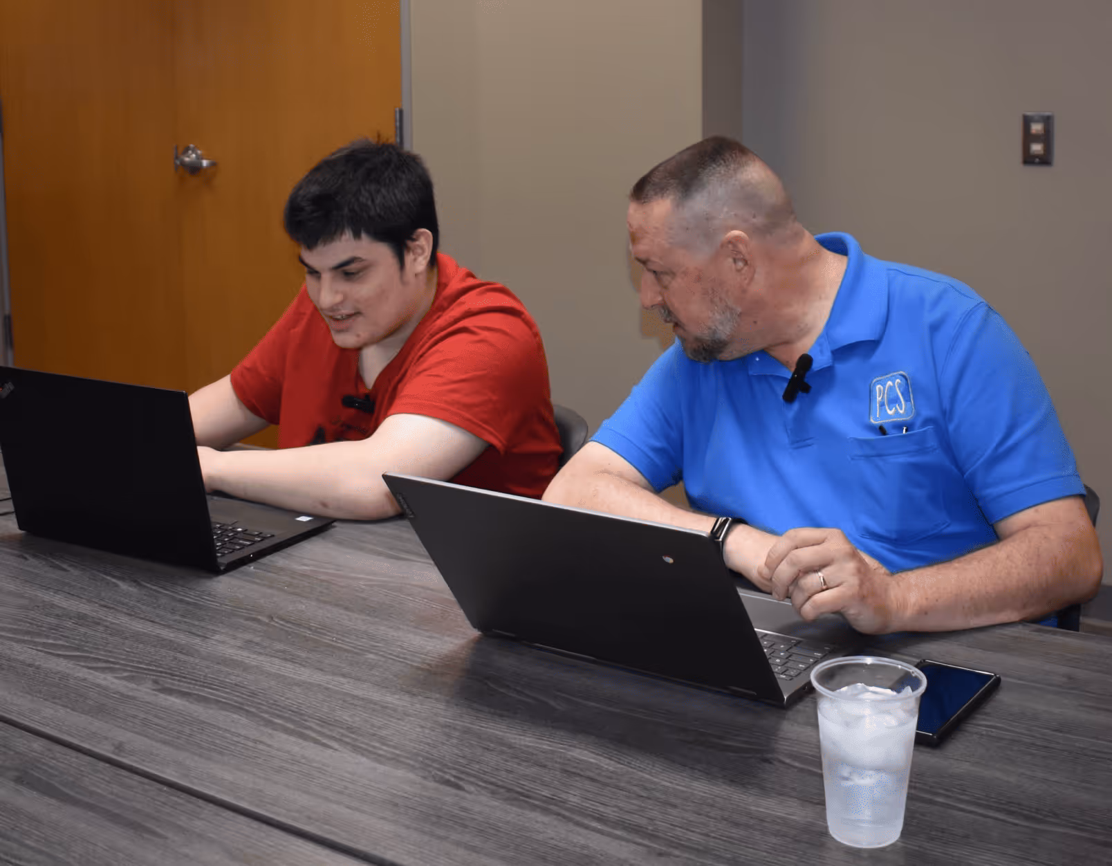 Two men sitting at a table working on laptops, one in a red shirt and the other in a blue polo shirt with a PCS logo.