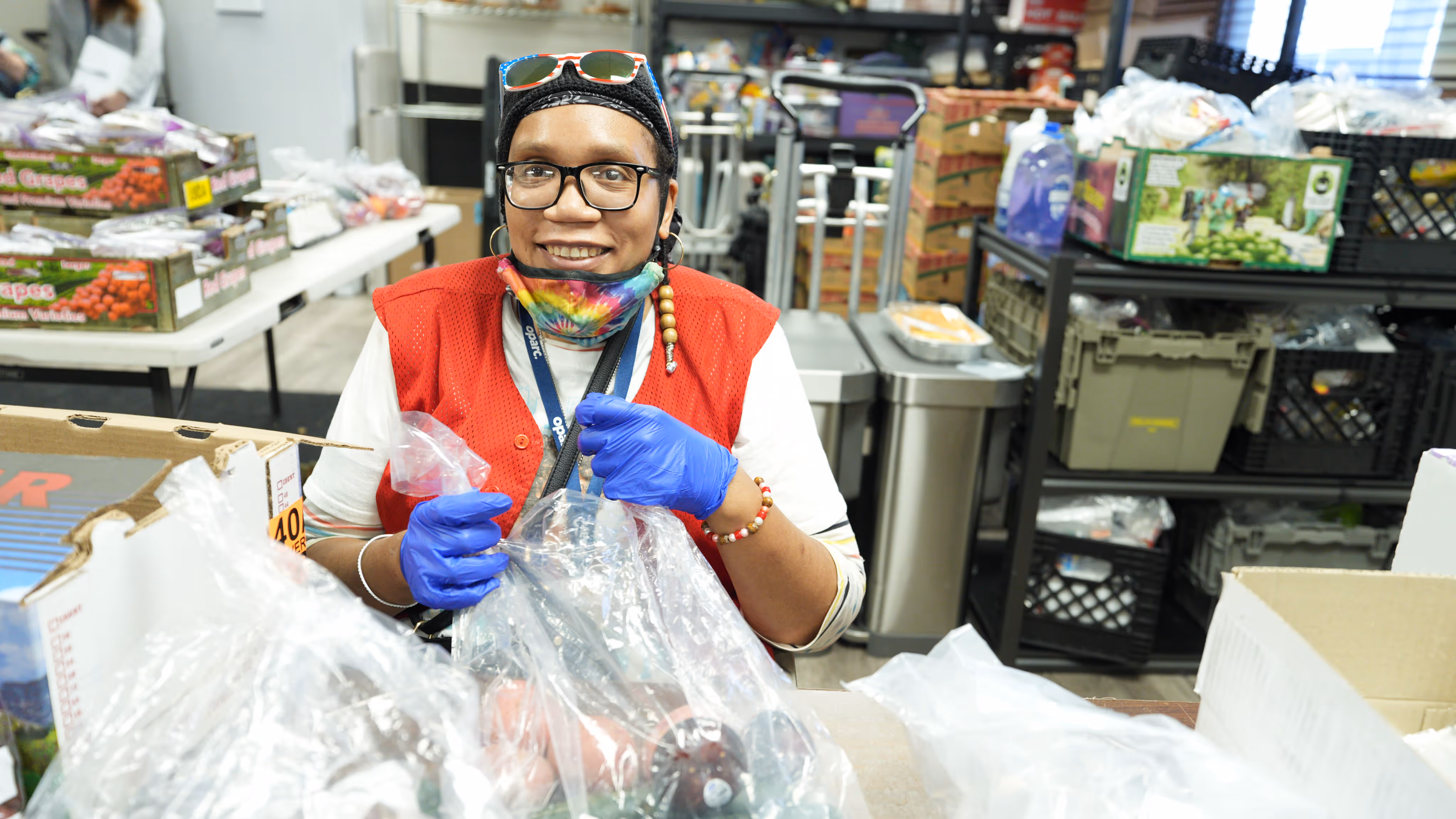 Smiling person wearing glasses, blue gloves, and a tie-dye mask around their neck, handling plastic bags in a room with food supplies.