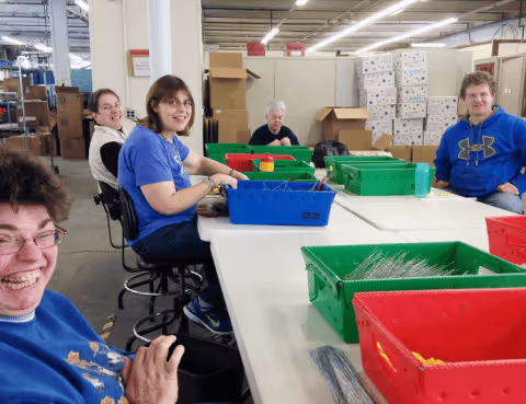 Five people sitting around a table in a warehouse, smiling and working with plastic bins filled with materials.