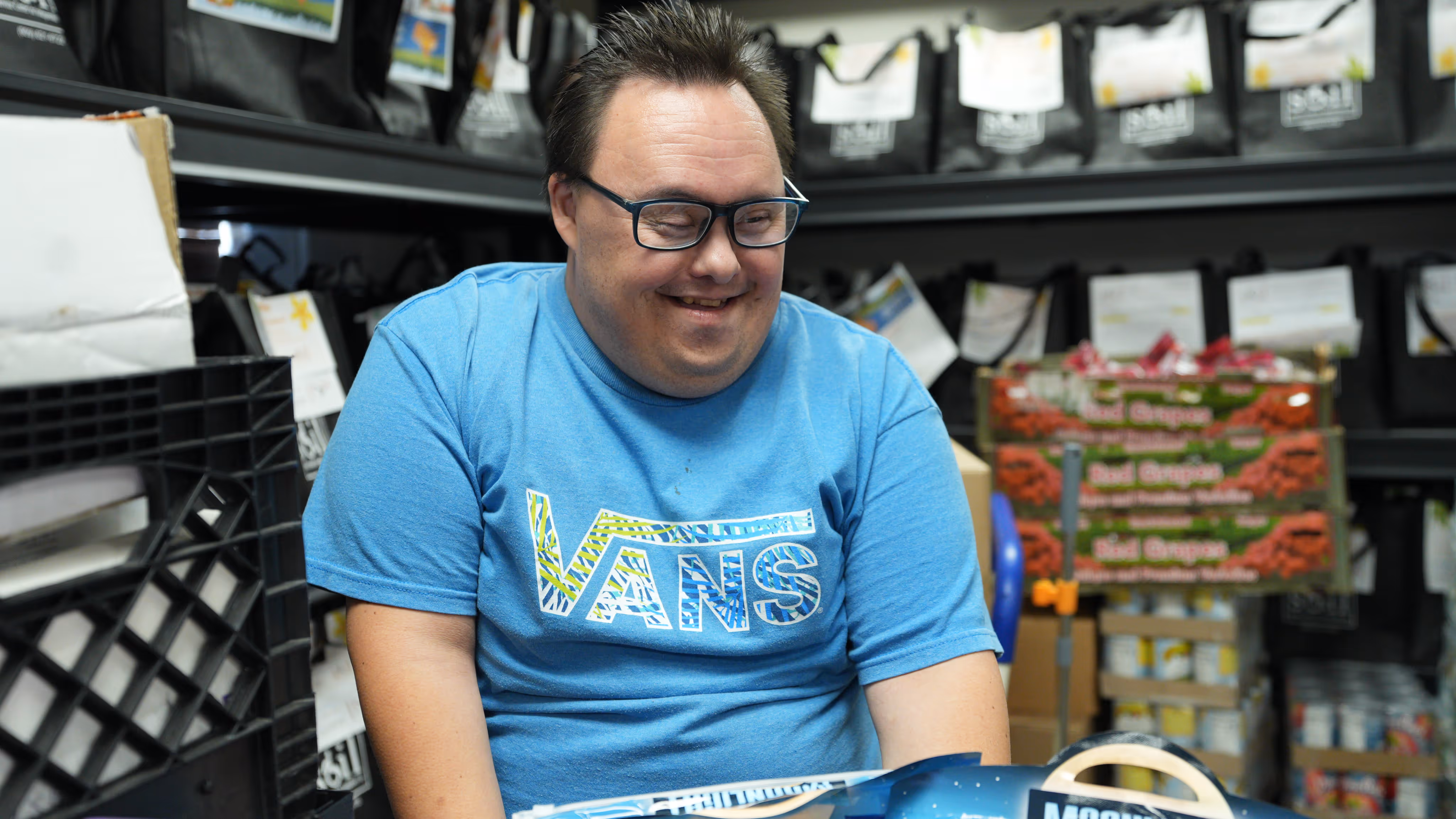 Smiling man wearing glasses and a blue Vans t-shirt sitting indoors with shelves of packaged goods in the background.