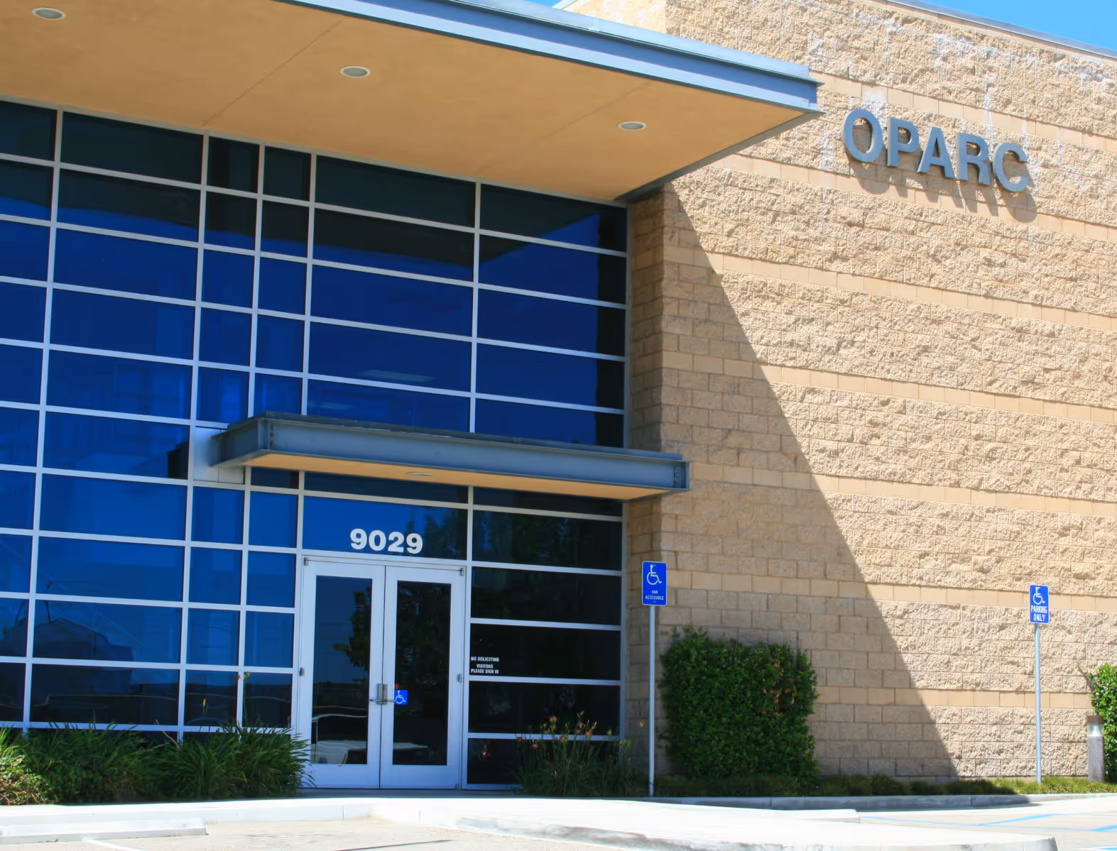 Building entrance with large blue-tinted windows, double glass doors numbered 9029, and a sign reading OPARC on a beige brick wall.