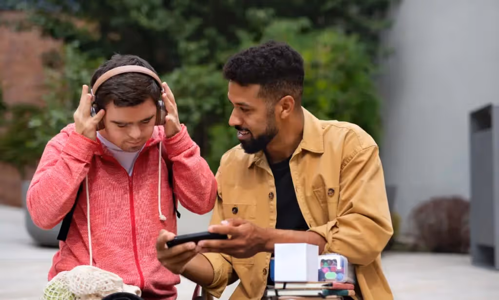 Two young men outdoors, one wearing headphones and a red jacket, the other showing something on a smartphone.