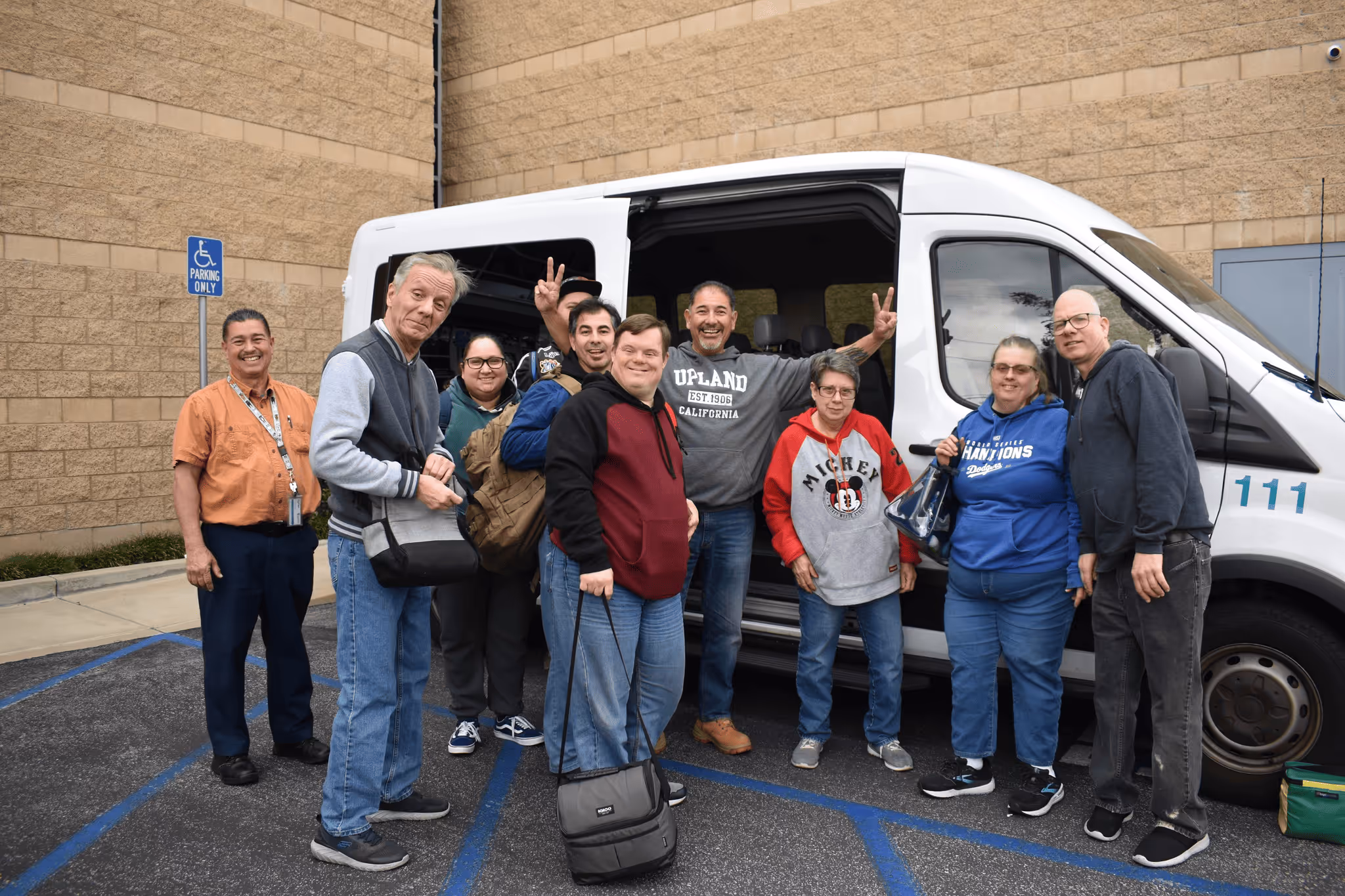 Group of ten diverse adults smiling and posing together beside a white van in a parking lot with a brick wall background.