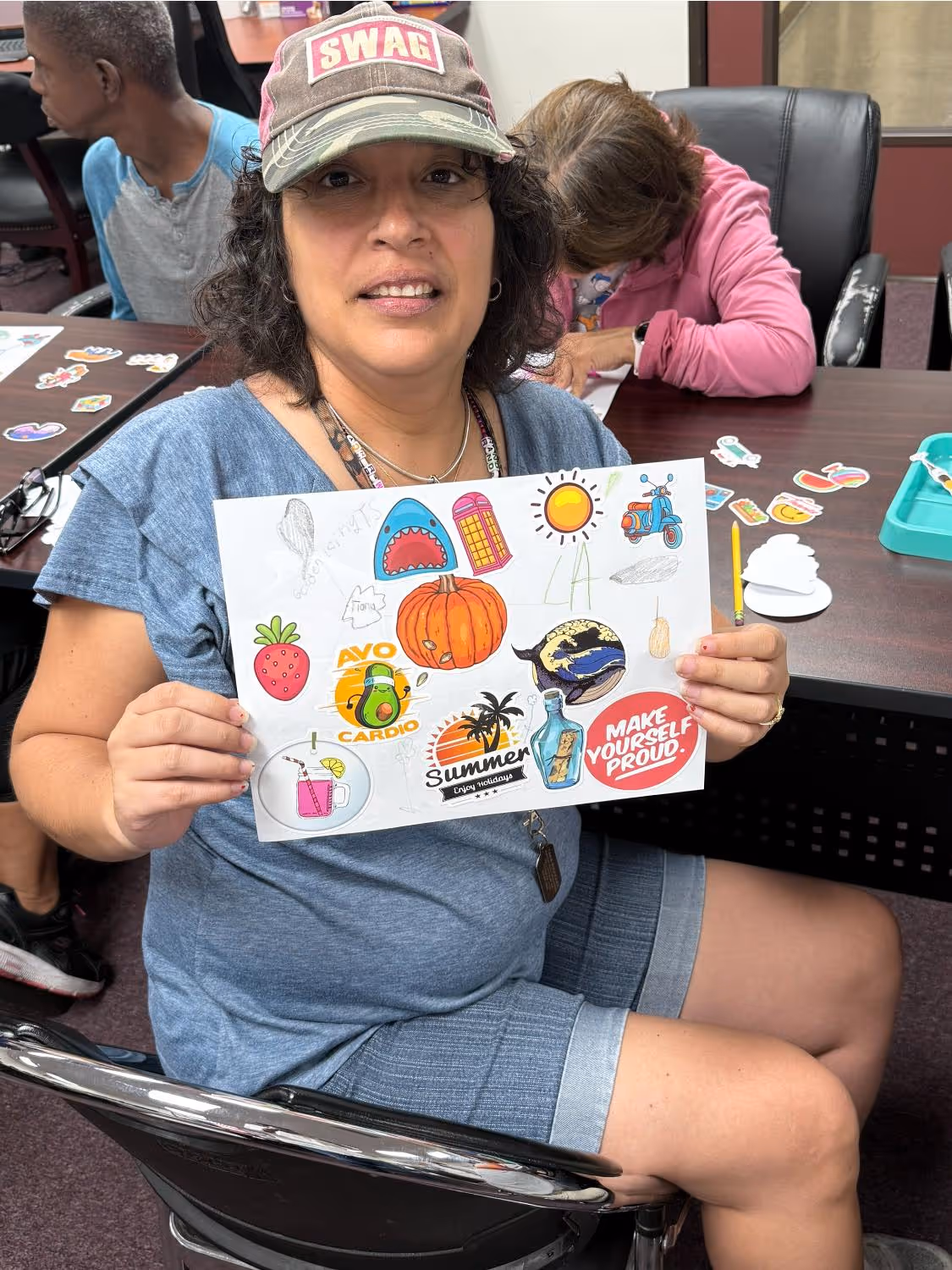 Smiling woman wearing a camo 'SWAG' cap holding a paper with colorful stickers including a pumpkin, sun, strawberry, scooter, and text 'MAKE YOURSELF PROUD.'