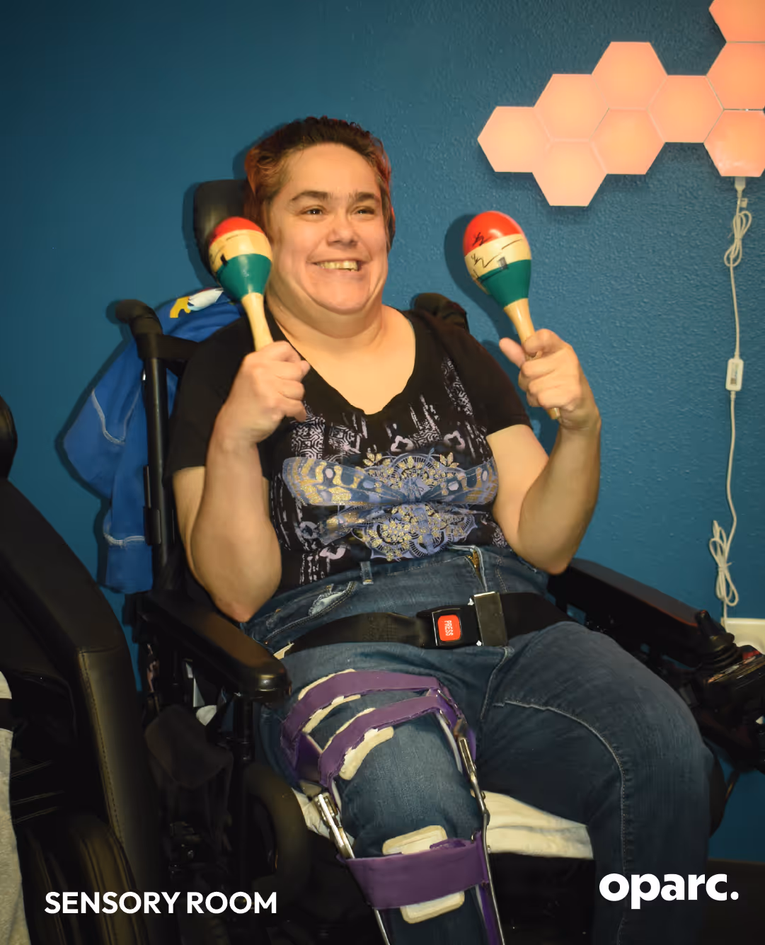 Smiling person in wheelchair holding colorful maracas in a sensory room with blue walls and hexagonal lights.