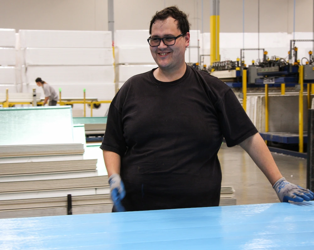 Smiling man wearing glasses and gloves standing by a blue sheet in an industrial warehouse.