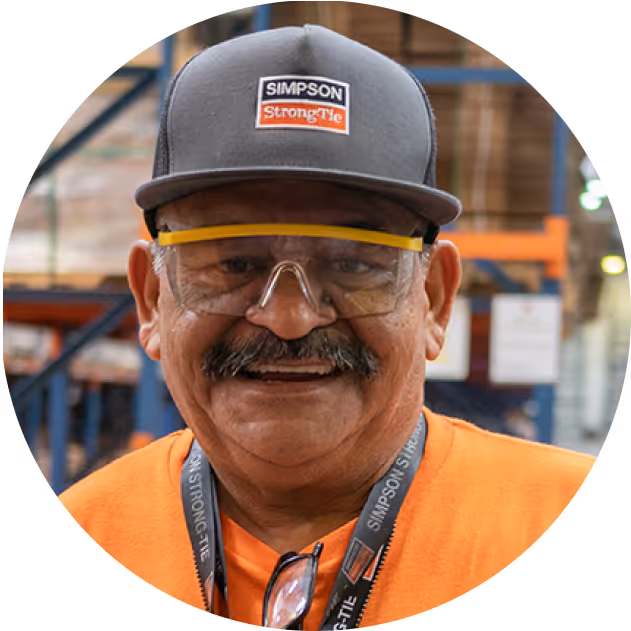 Smiling man wearing safety glasses, a gray Simpson Strong-Tie cap, and an orange shirt inside a warehouse.