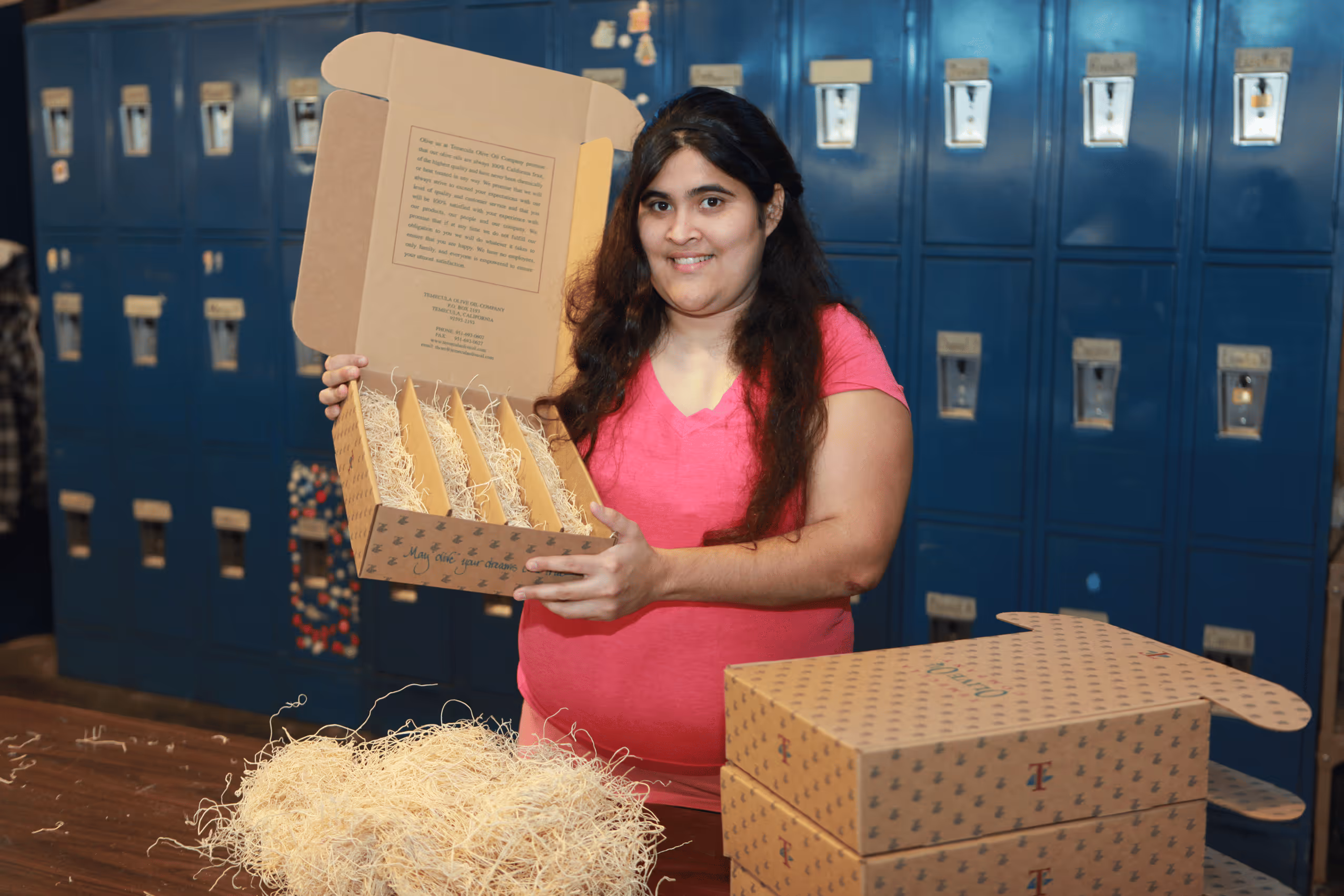 Woman in a pink shirt holding an open cardboard box with compartments and packing material, standing in front of blue lockers.