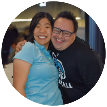 Two smiling people embracing each other, one wearing a light blue shirt and the other wearing glasses and a black shirt.