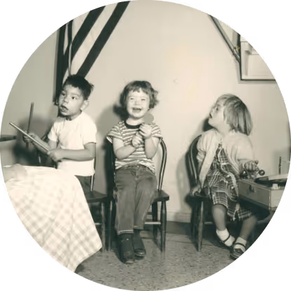 Three young children sitting on chairs indoors, with one child happily clapping in the middle.