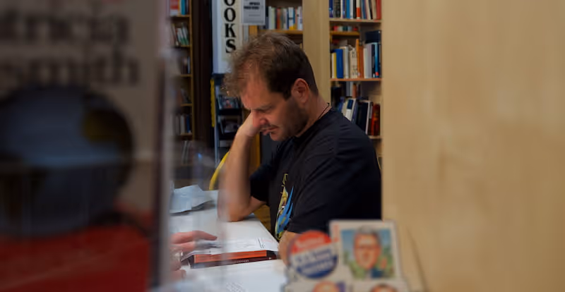 Man sitting at a table in a bookstore reading a book with bookshelves in the background.