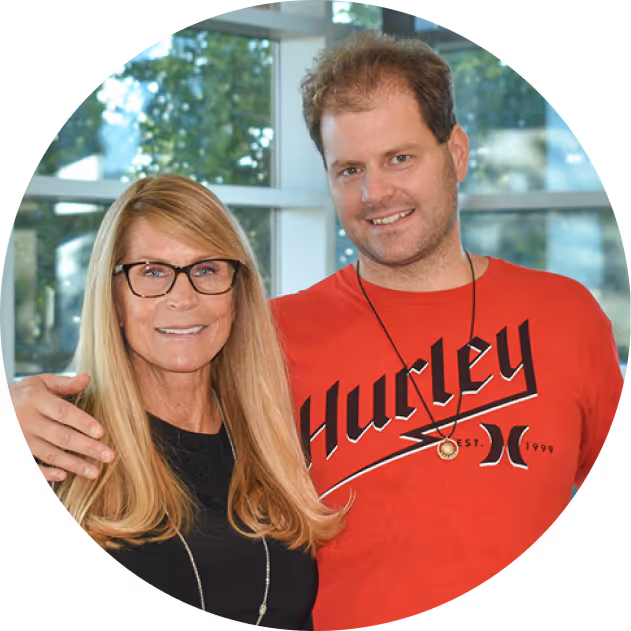 Smiling man in a red shirt with arm around his smiling mother with long blonde hair and glasses, indoors by windows.