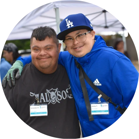 Two smiling men with Down syndrome hugging outdoors under a white tent, wearing name badges.