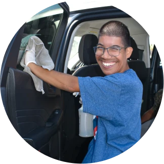 Smiling person wearing glasses and a blue shirt cleaning a car door with a cloth.