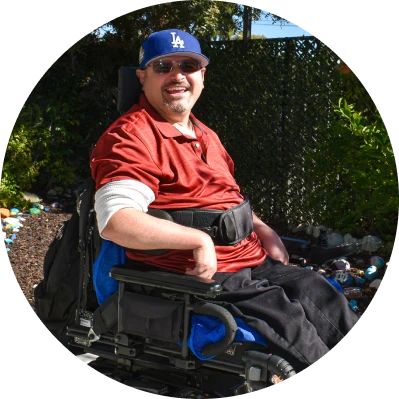 Smiling man in a motorized wheelchair outdoors wearing a blue Los Angeles Dodgers cap and sunglasses.