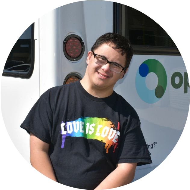 Smiling person wearing glasses and a black 'Love is Love' rainbow t-shirt standing in front of a vehicle.