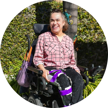 Smiling woman sitting in a motorized wheelchair outdoors with greenery and a tree in the background.