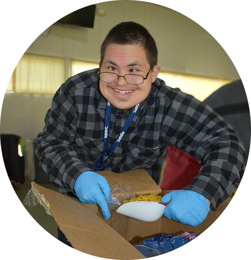 Smiling person wearing glasses and blue gloves packing food into a box indoors.