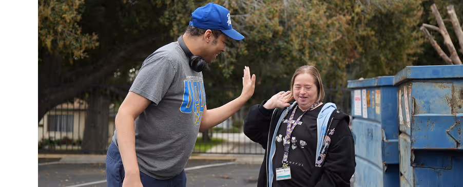 Two people smiling and giving each other a high five outdoors near blue dumpsters.