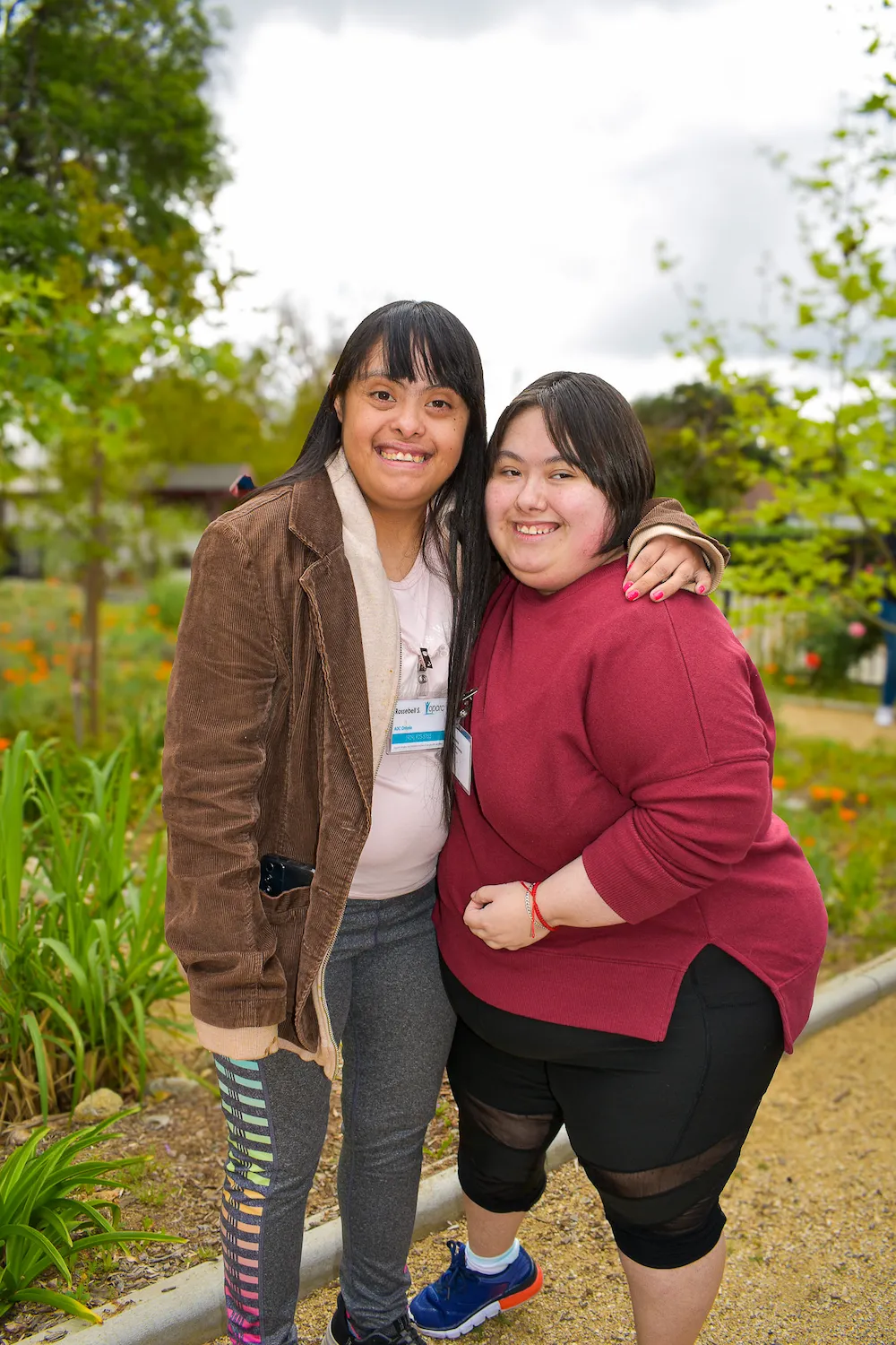 Two smiling young women standing outdoors with one arm around each other, surrounded by greenery.