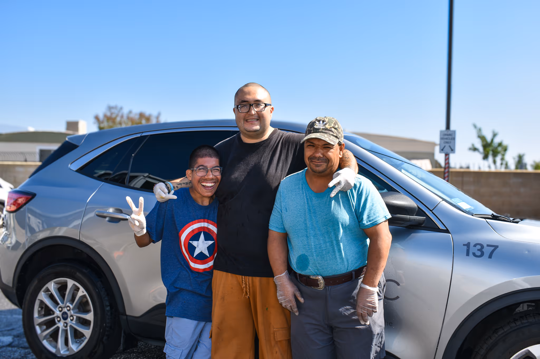 Three men wearing gloves smiling and posing in front of a silver car under clear sky.