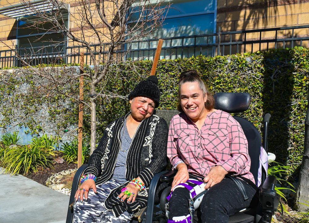 Two women sitting outside in the sun, smiling; one wearing a black knit hat and patterned cardigan, the other in a pink plaid shirt and sitting in a wheelchair with a leg brace.
