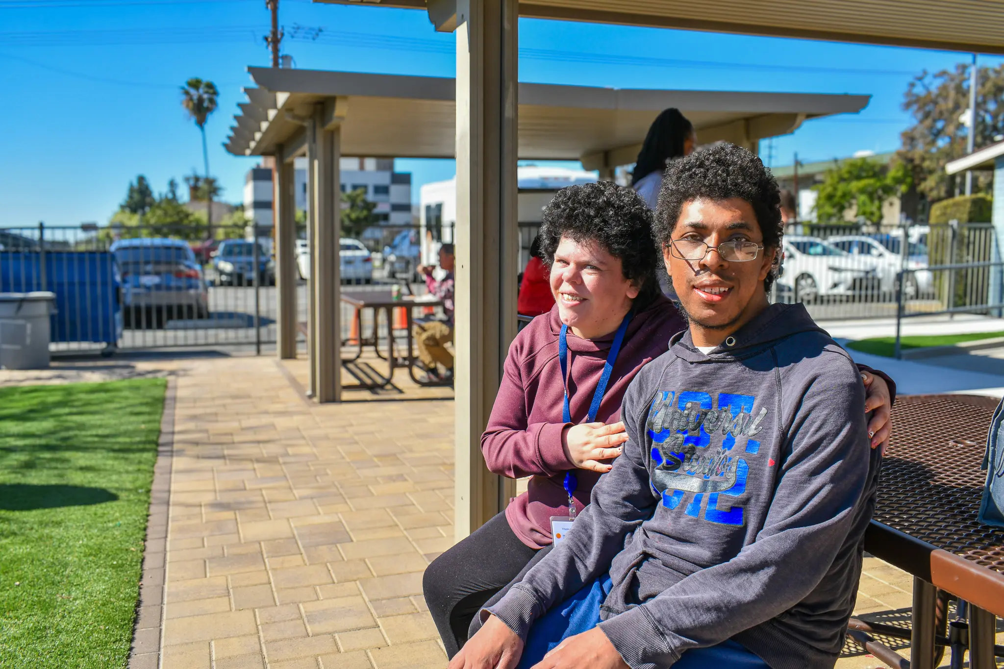 Two people sitting closely on a bench outdoors under a shaded area on a sunny day, smiling towards the camera.