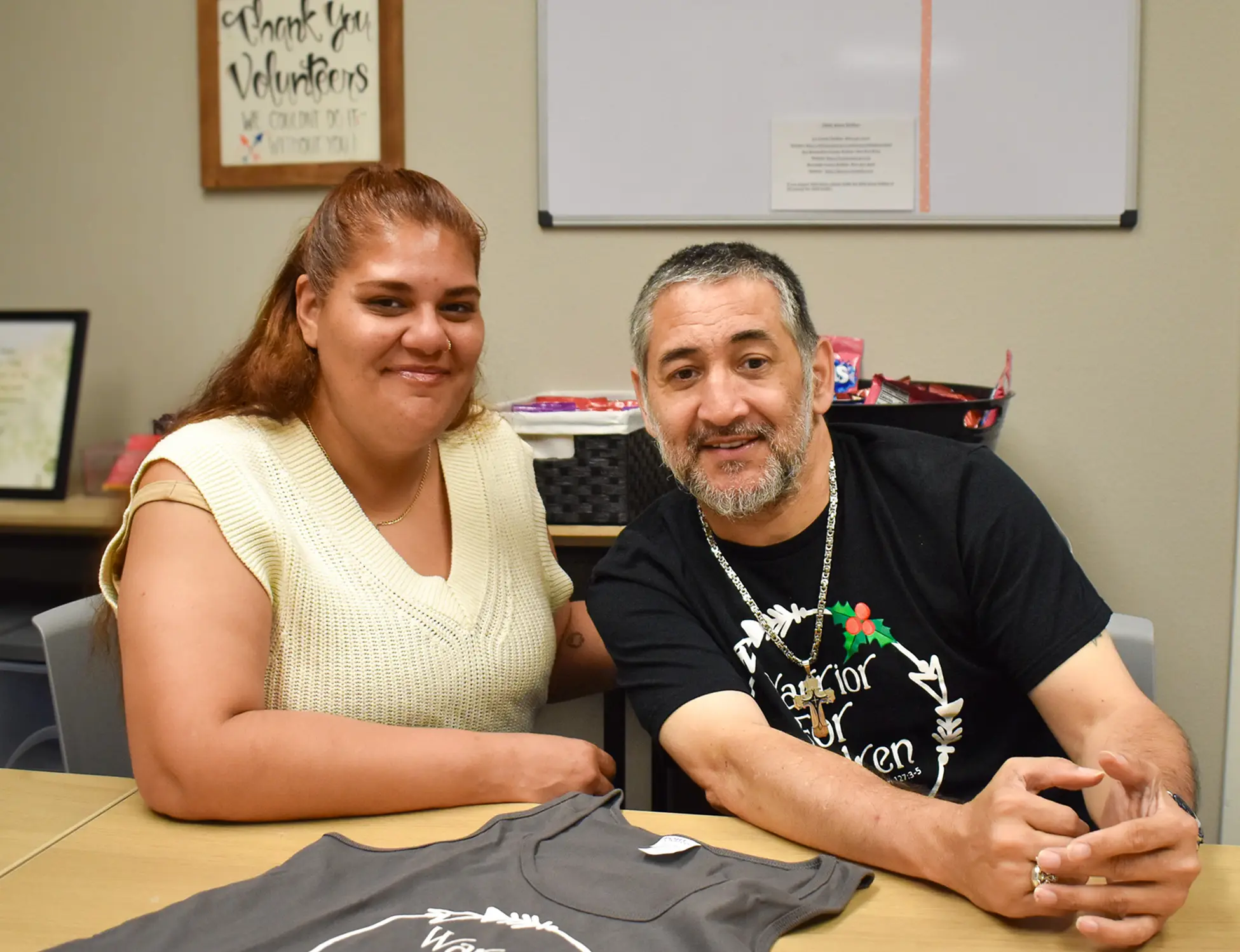 Two people smiling and sitting at a table with a folded T-shirt in front of them in an office setting.