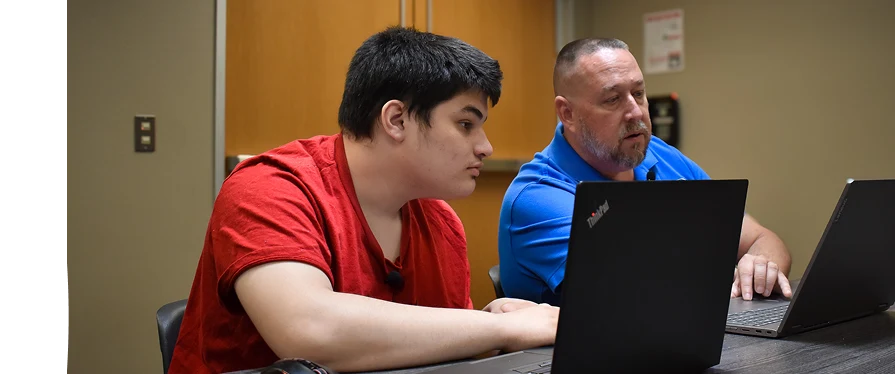 A young man in a red shirt and an older man in a blue shirt sit at a table working on laptops together.
