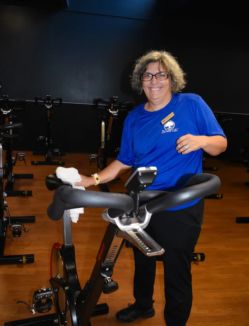 Smiling woman wearing glasses and a blue shirt with a name tag standing next to a stationary exercise bike in a gym.