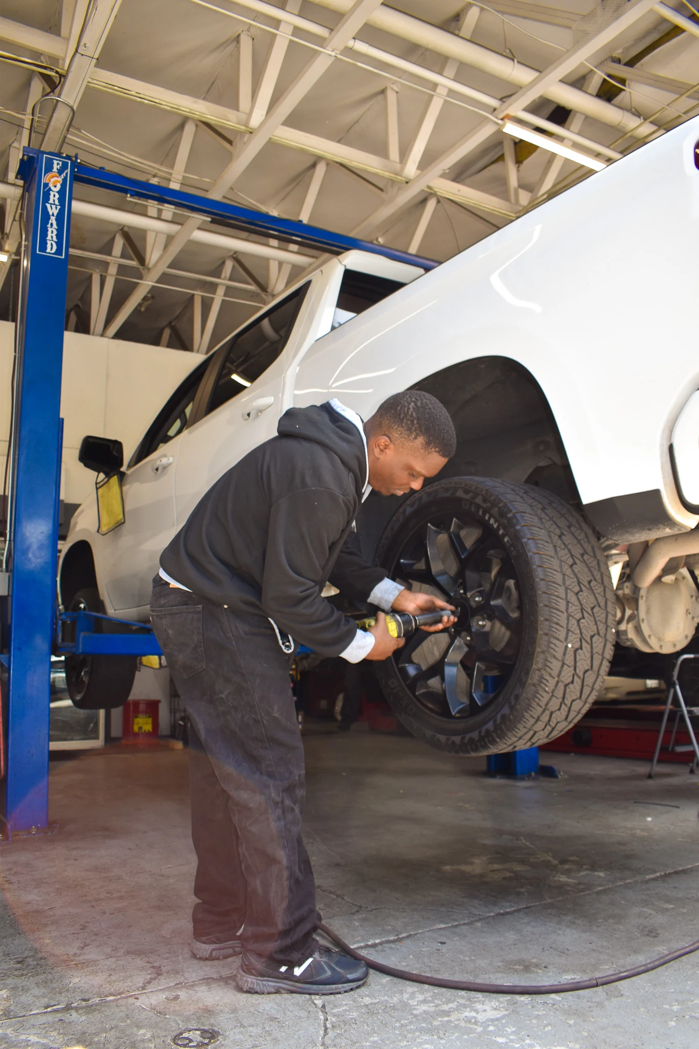 Mechanic using a power tool to work on the rear wheel of a white pickup truck lifted in a garage.