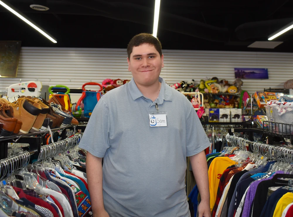 Smiling man wearing a name tag standing between clothing racks in a store filled with shoes and toys.