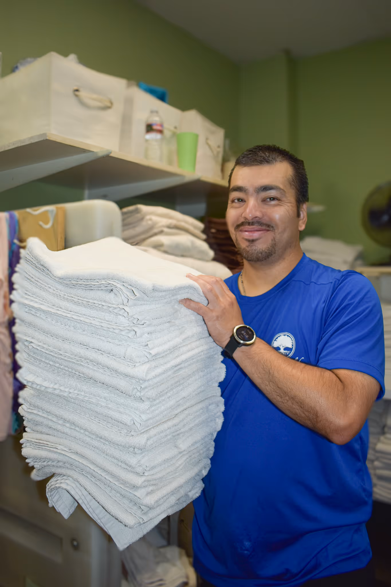 Man in blue shirt smiling and holding a large stack of folded white towels in a laundry room.