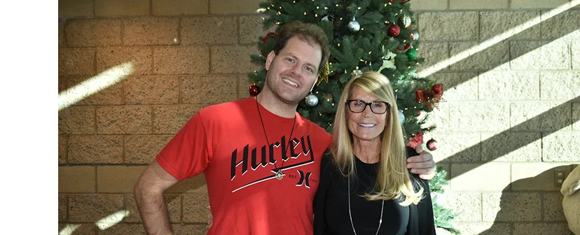 Man in a red t-shirt smiling with an arm around his mom with long blonde hair and glasses, standing in front of a decorated Christmas tree.