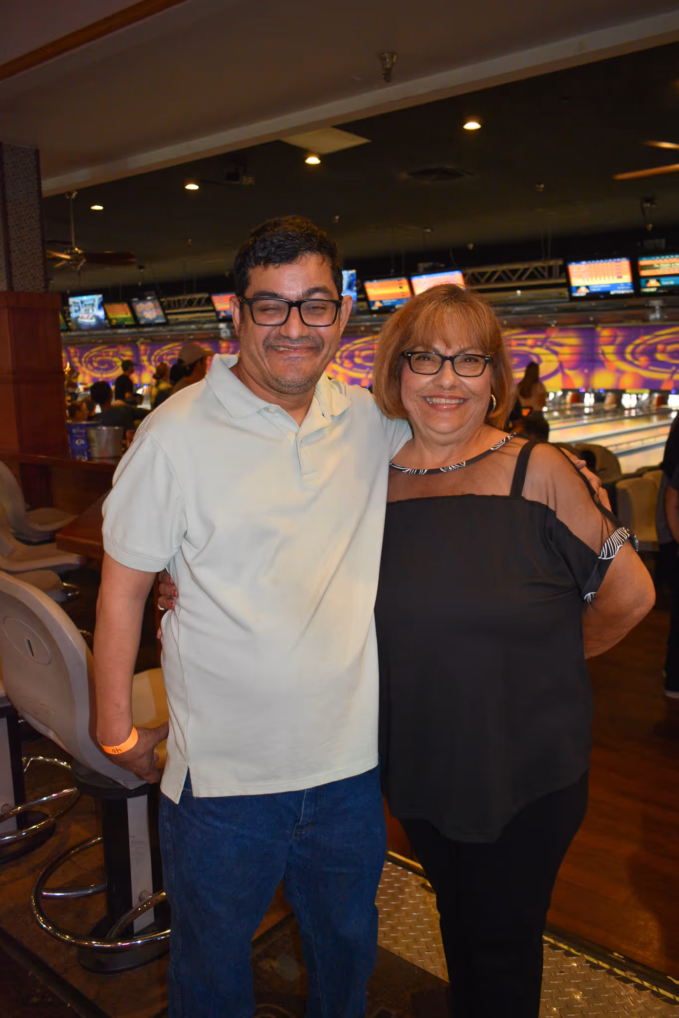 Smiling man and woman wearing glasses, standing close together in a bowling alley.