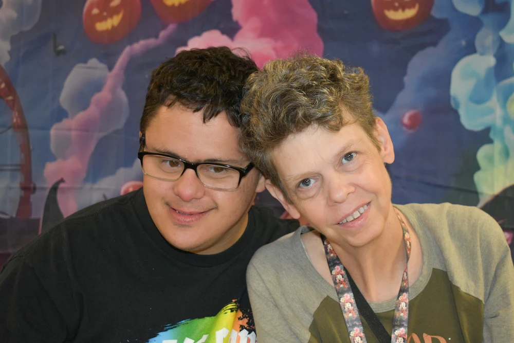 Smiling young man with glasses and woman posing closely in front of a colorful Halloween-themed backdrop.