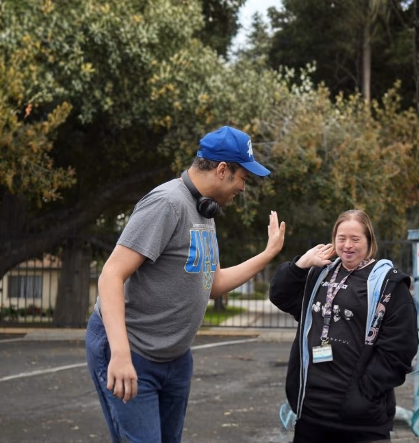 Smiling young man giving a high five to a woman with Down syndrome near blue dumpsters in an outdoor parking area.