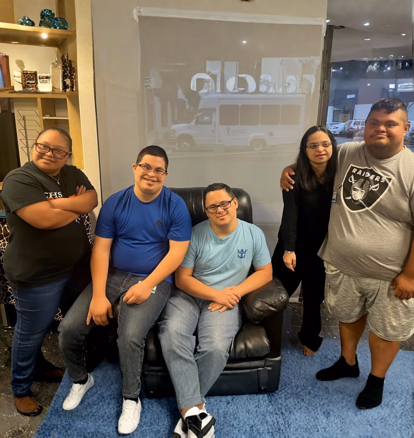 Five people with Down syndrome smiling together indoors, some seated on a black armchair and others standing nearby.