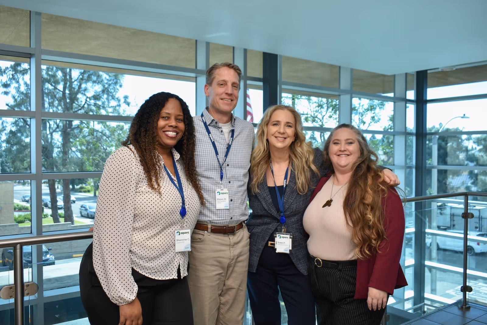 Four smiling diverse colleagues posing together inside a modern office building with large windows.