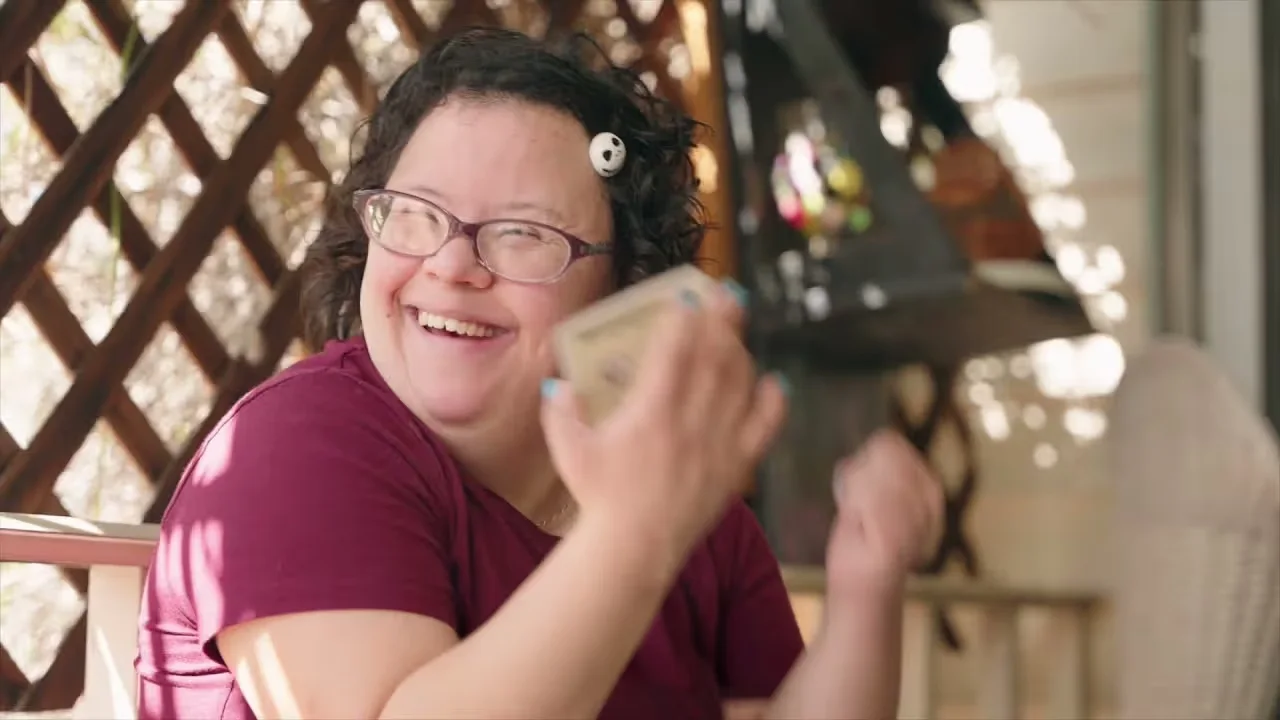 Smiling woman with glasses and curly hair holding a ten-dollar bill, sitting outdoors with wooden lattice background.