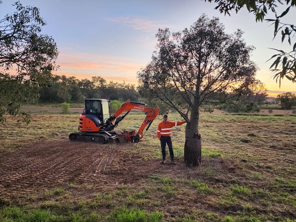 Man in hi-vis gear stands by a tree next to an orange excavator in a cleared field at sunset.