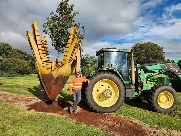 Man in hi-vis leans on tractor beside a tree spade holding a tree above a fresh dirt hole.