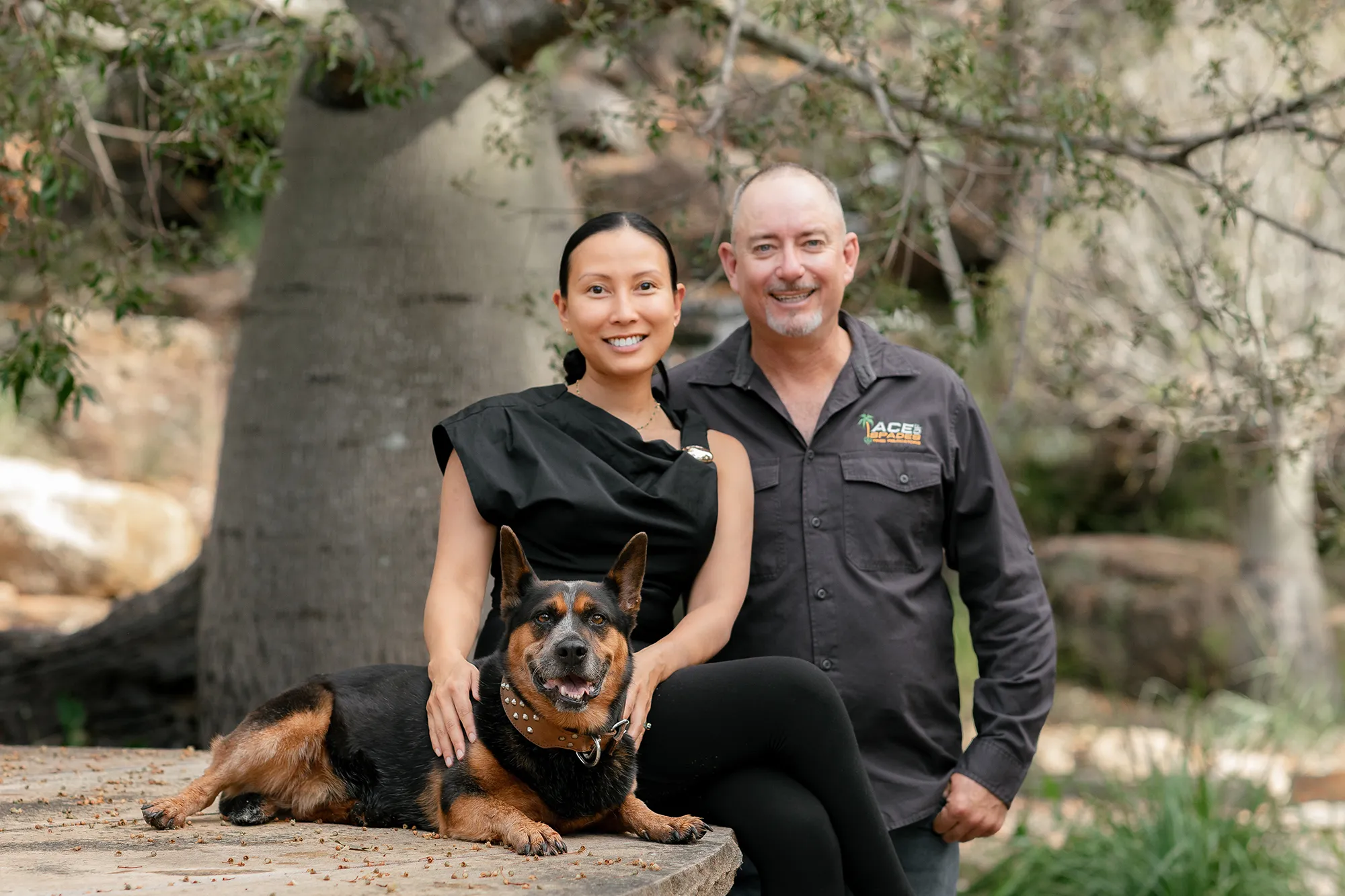 Woman seated on a stone slab with a black-and-tan dog lying in front, while a man stands beside her; all smiling in a wooded outdoor setting.