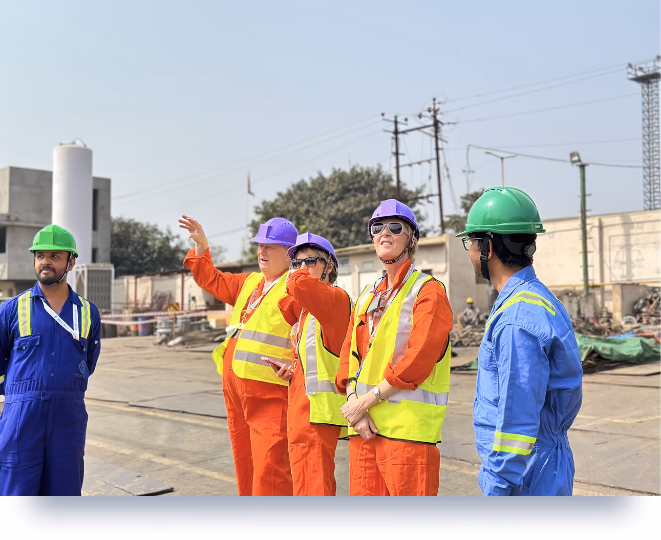 Five industrial workers in safety gear, hard hats, and vests at an outdoor industrial site.