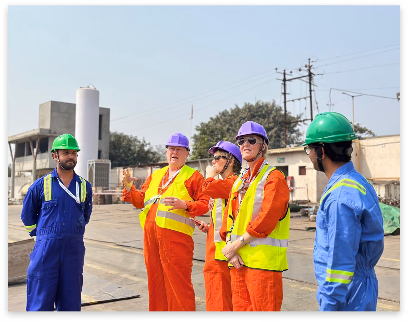 Five construction workers in safety gear and helmets having a discussion outdoors on site.